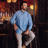 A man in a Tom Beckbe Hanover Chambray Shirt and beige pants sits on a bar stool in a dimly lit bar.