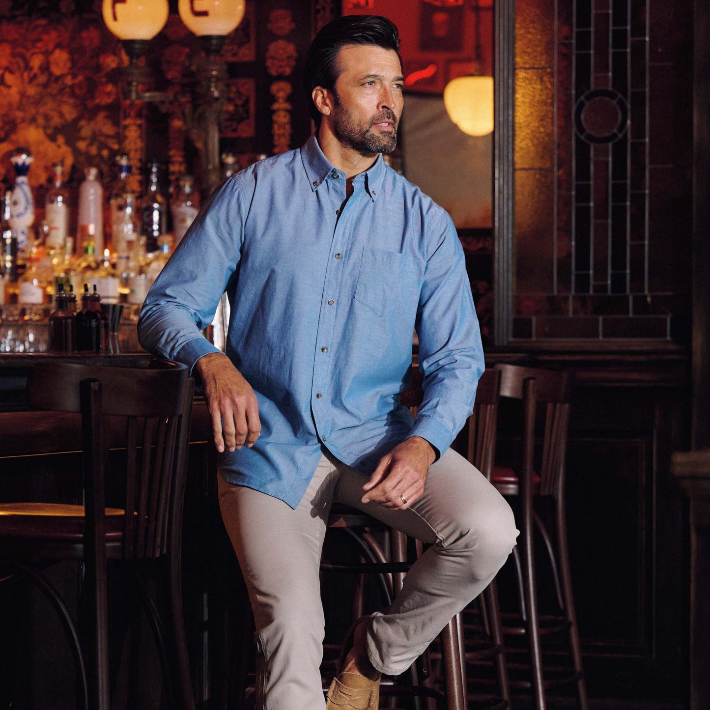 A man in a Tom Beckbe Hanover Chambray Shirt and beige pants sits on a bar stool in a dimly lit bar.
