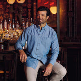 A man in a Tom Beckbe Hanover Chambray Shirt and beige pants sits at a bar with bottles and dim lighting behind him.