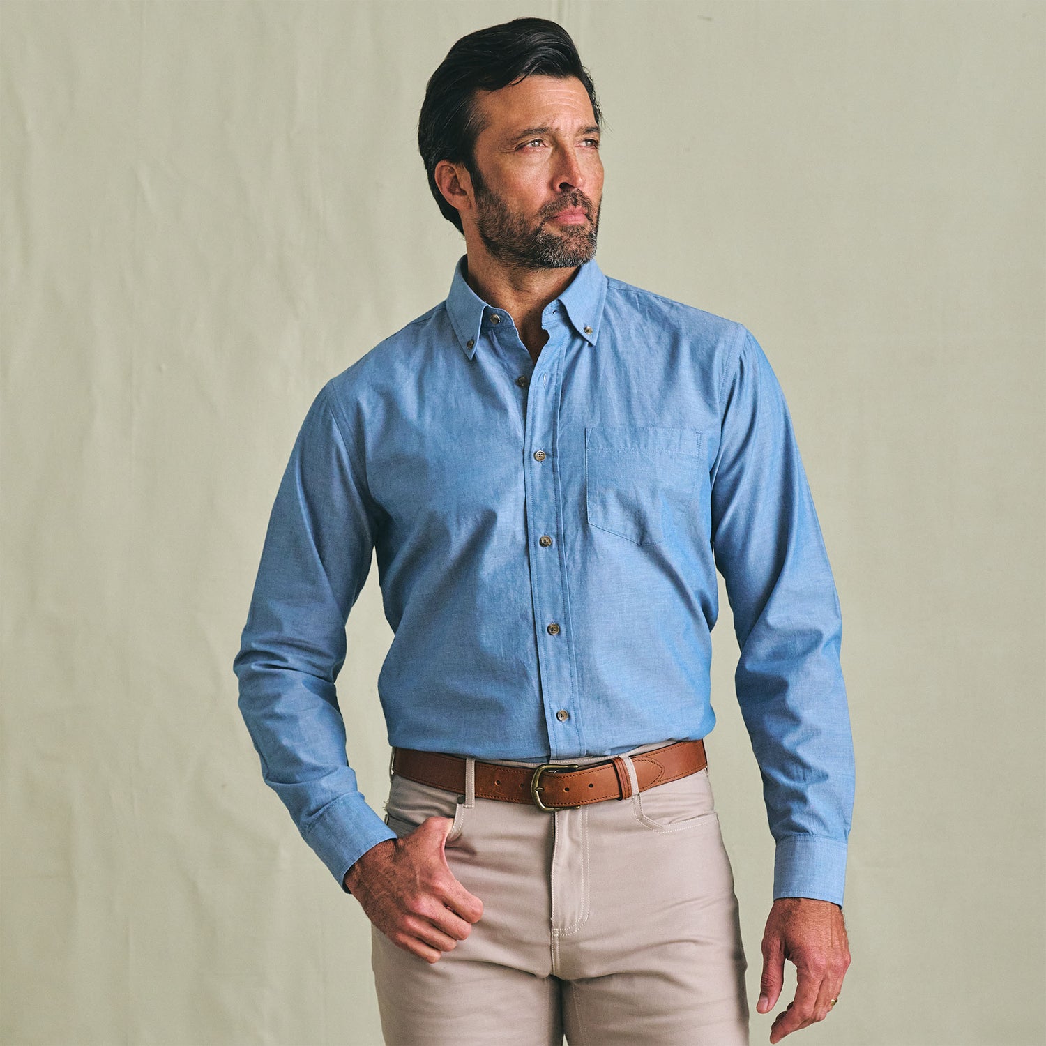 Man wears the Tom Beckbe Hanover Chambray Shirt and beige pants, standing against a plain light background, looking aside.
