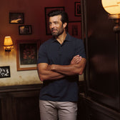 Bearded man indoors, arms crossed, wears Tom Beckbe Hutton Performance Polo and beige pants, surrounded by framed art.