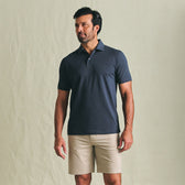 Man wears Tom Beckbe Hutton Performance Polo and light shorts against a plain, light-colored background.