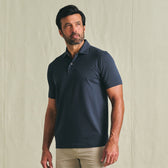 Man wearing a Tom Beckbe Hutton Performance Polo in dark navy and beige pants, standing against a light background.
