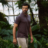 A man in a Tom Beckbe Hutton Performance Polo and beige shorts stands among lush green plants in a garden setting.