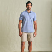 Man wearing the Tom Beckbe Hutton Performance Polo in light blue stripes with beige shorts against a plain background.