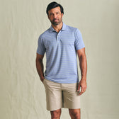 Man in a Tom Beckbe Hutton Performance Polo and beige shorts stands against a plain light background.