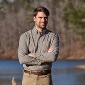 Man in Tom Beckbe Knapp Cotton Twill Shirt and khakis stands outdoors by water and trees, arms crossed.
