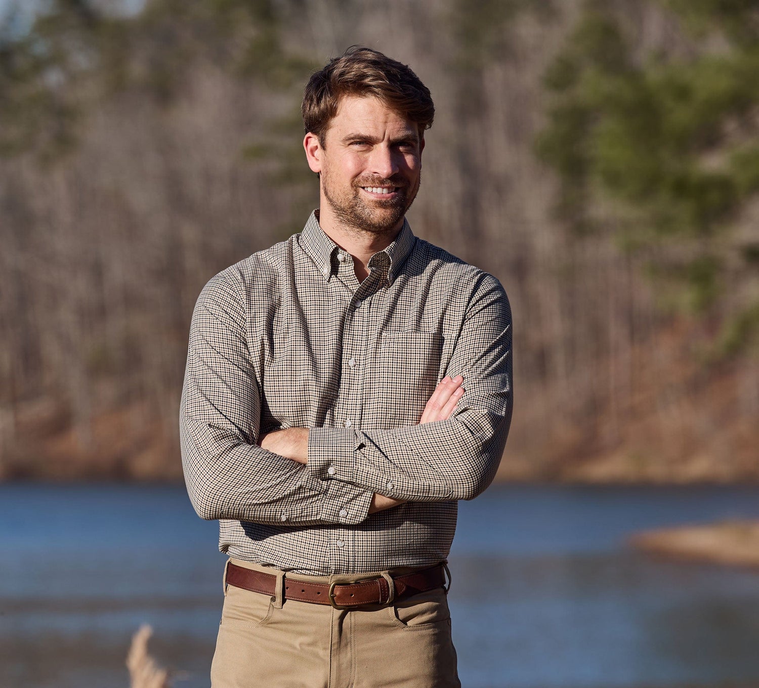 Man in Tom Beckbe Knapp Cotton Twill Shirt and khakis stands outdoors by water and trees, arms crossed.