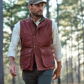 A man in a Tom Beckbe Fox Red Kinsman Vest, plaid shirt, and cap walks through a forest with trees behind him.