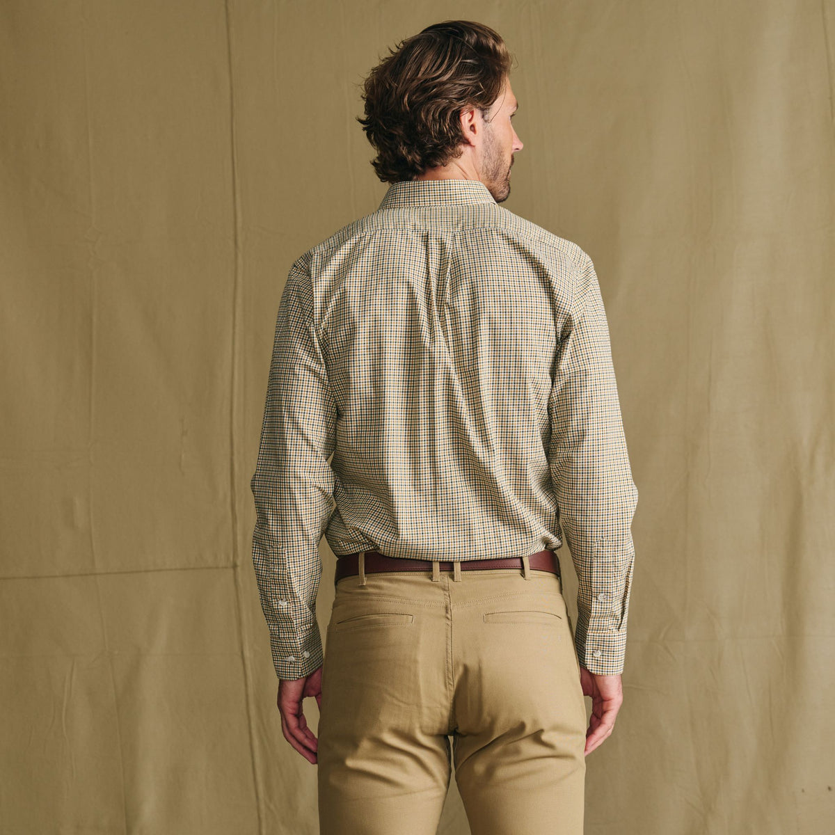 Man standing with his back to the camera, wearing a checked shirt and khaki pants against a plain beige background.