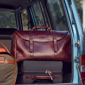 A Tom Beckbe Leather Weekender Duffel Bag sits among luggage in the trunk of a van.