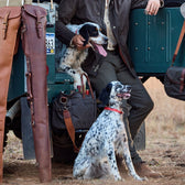 Two spotted dogs with red collars sit by an off-road vehicle, next to a person with a Tom Beckbe Leather Gun Sleeve.