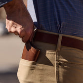 A person slips a Tom Beckbe Leather Passport Wallet into the back pocket of beige pants, blue checkered shirt tucked in.