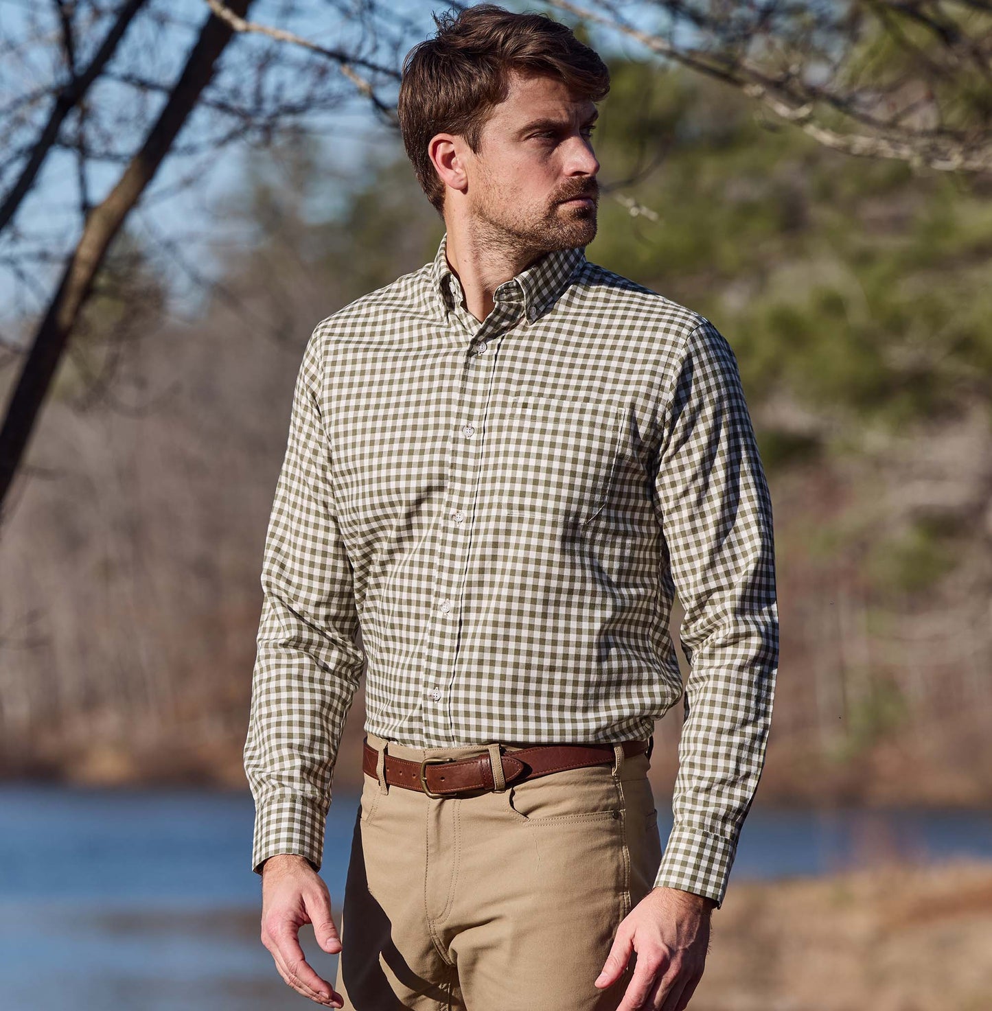 Man wears a Tom Beckbe Knapp Cotton Twill Shirt and khaki pants outdoors by water and trees.
