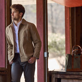 Man in a Livingston Jacket and gray pants stands by a red wooden structure, looking outside. A green bag is on a bench nearby.