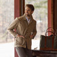 A man in a Livingston Jacket by Tom Beckbe smiles with hands in pockets, leaning against a wooden post on a porch. A green and brown tote bag is on the table next to him, while blurred trees and daylight in the background capture outdoor utility.