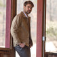 A bearded man leans against a rustic wooden structure, wearing the Livingston Jacket by Tom Beckbe in light brown over a white shirt and dark pants. He gazes to the side, surrounded by blurred hints of trees and natural surroundings.