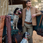 A man wearing a Tom Beckbe Logan Bomber Jacket leans on a jeep with dogs, amidst leather bags in a vintage setting.