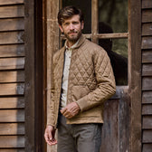 A man in a vintage-style Logan Bomber Jacket by Tom Beckbe and gray pants stands by a wooden building, facing the camera.