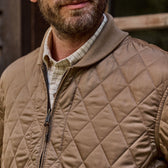 Bearded man outdoors, wearing a tan Logan Bomber Jacket by Tom Beckbe over a plaid shirt, near a wooden window.