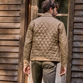 A man in a Tom Beckbe Logan Bomber Jacket and green pants stands outside a wooden building, facing away from the camera.