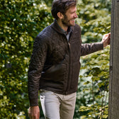 Man in a vintage-style Tom Beckbe Logan Bomber Jacket and beige pants smiles near wooden structure with greenery.