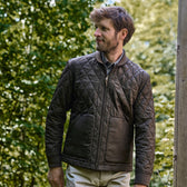 Man in a Tom Beckbe Logan Bomber Jacket stands outside, quilted vintage-style brown with green trees behind him.
