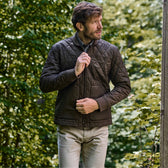 Man in Tom Beckbes Logan Bomber Jacket adjusts collar, embracing vintage style among green foliage.