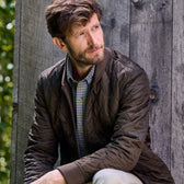 Man in a brown Tom Beckbe Logan Bomber Jacket and plaid shirt, sitting against a wooden wall, looking to the side.