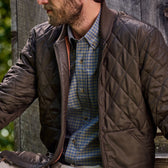 Man in a Tom Beckbe Logan Bomber Jacket over a blue checkered shirt, sitting outside near a wooden wall.