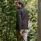 Man in a brown Tom Beckbe Logan Bomber Jacket and beige pants outdoors, touching his back near green foliage.
