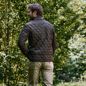 A man in a Tom Beckbe Logan Bomber Jacket and beige pants stands facing away in a forest.