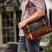 Man in checkered shirt carries a Tom Beckbe Messenger Bag and suitcase outdoors.