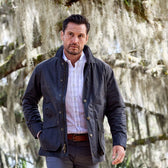 A man in a classic fit dark jacket and a Tom Beckbe Monroe Poplin Shirt walks past moss-covered trees outdoors.