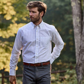 A man in a Tom Beckbe Monroe Poplin Shirt and gray pants stands near a tree, surrounded by lush greenery.