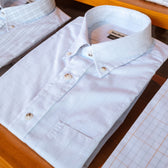 The Monroe Poplin Shirt by Tom Beckbe in light blue plaid is neatly folded on a wooden surface among other shirts.