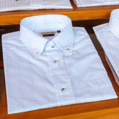 The Monroe Poplin Shirt by Tom Beckbe, featuring a subtle grid pattern, is neatly folded on a wooden shelf.