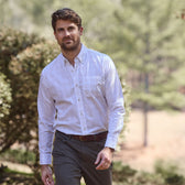 A man wearing a Tom Beckbe Monroe Poplin Shirt and dark pants strolls outdoors amid a serene backdrop of trees.