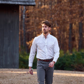 A man in a Tom Beckbe Monroe Poplin Shirt and gray pants walks outside near a wooden building and autumn trees.