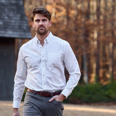 A man in a Tom Beckbe Monroe Poplin Shirt and gray pants walks outdoors near trees and a building.