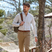 Man strolling in a Monroe Poplin Shirt by Tom Beckbe and tan pants, backpack on, trees and a hammock behind him.
