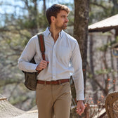 A man with a backpack walks outdoors in a light blue Monroe Poplin Shirt by Tom Beckbe and khaki pants, with trees behind.
