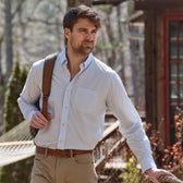 A man in a Tom Beckbe Monroe Poplin Shirt and khaki pants stands outside with a backpack strap over his shoulder.