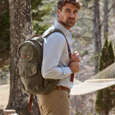 A man in a Tom Beckbe Monroe Poplin Shirt, khakis, and a green backpack stands by a hammock in the forest.