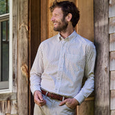 Man in a Monroe Poplin Shirt by Tom Beckbe leans against a wooden wall, smiling and looking to the side outdoors.