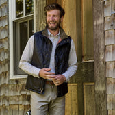 A man in a Tom Beckbe Monroe Poplin Shirt and dark vest smiles by a wooden door with shingle siding.