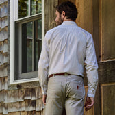 Man in a Monroe Poplin Shirt by Tom Beckbe and khaki pants stands by a wooden house, facing away.