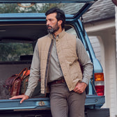 Man in a beige vest and gray pants leans on SUV, luggage inside, wearing Tom Beckbe Monroe Poplin Shirt.
