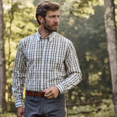 A man in a classic Monroe Poplin Shirt by Tom Beckbe stands among trees, looking right, surrounded by lush greenery.
