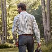 A person in a Monroe Poplin Shirt and gray pants from Tom Beckbe strolls through a wooded area with trees in the background.